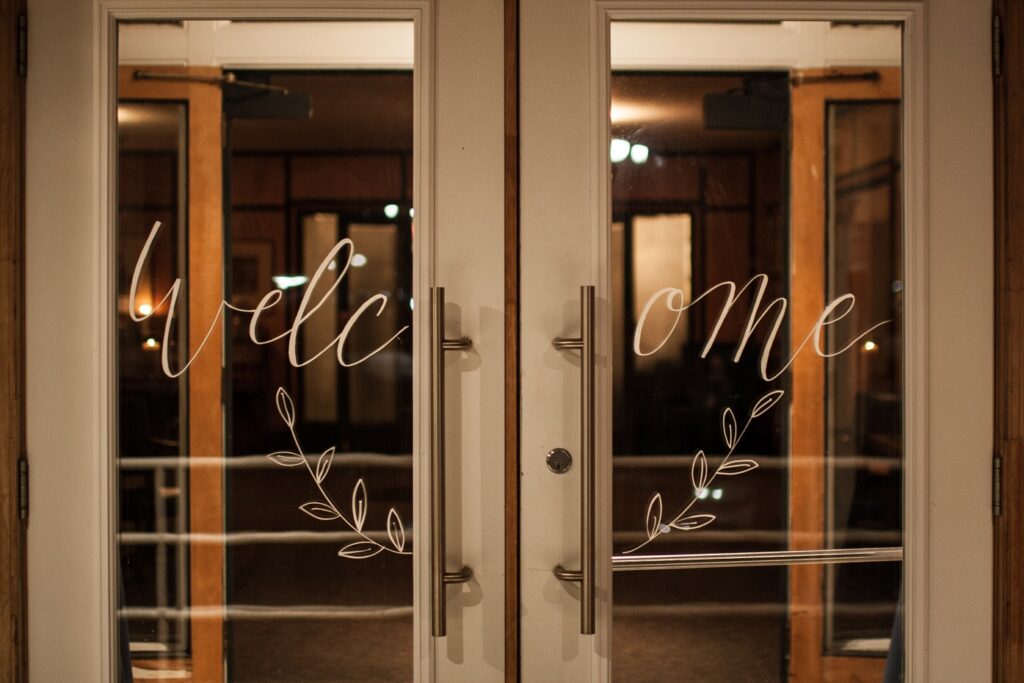 Glass double doors with “Welcome” written in cursive across both panels, along with simple leaf designs underneath the text, invite guests into the Henley Room. Warm indoor lighting is visible through the glass.