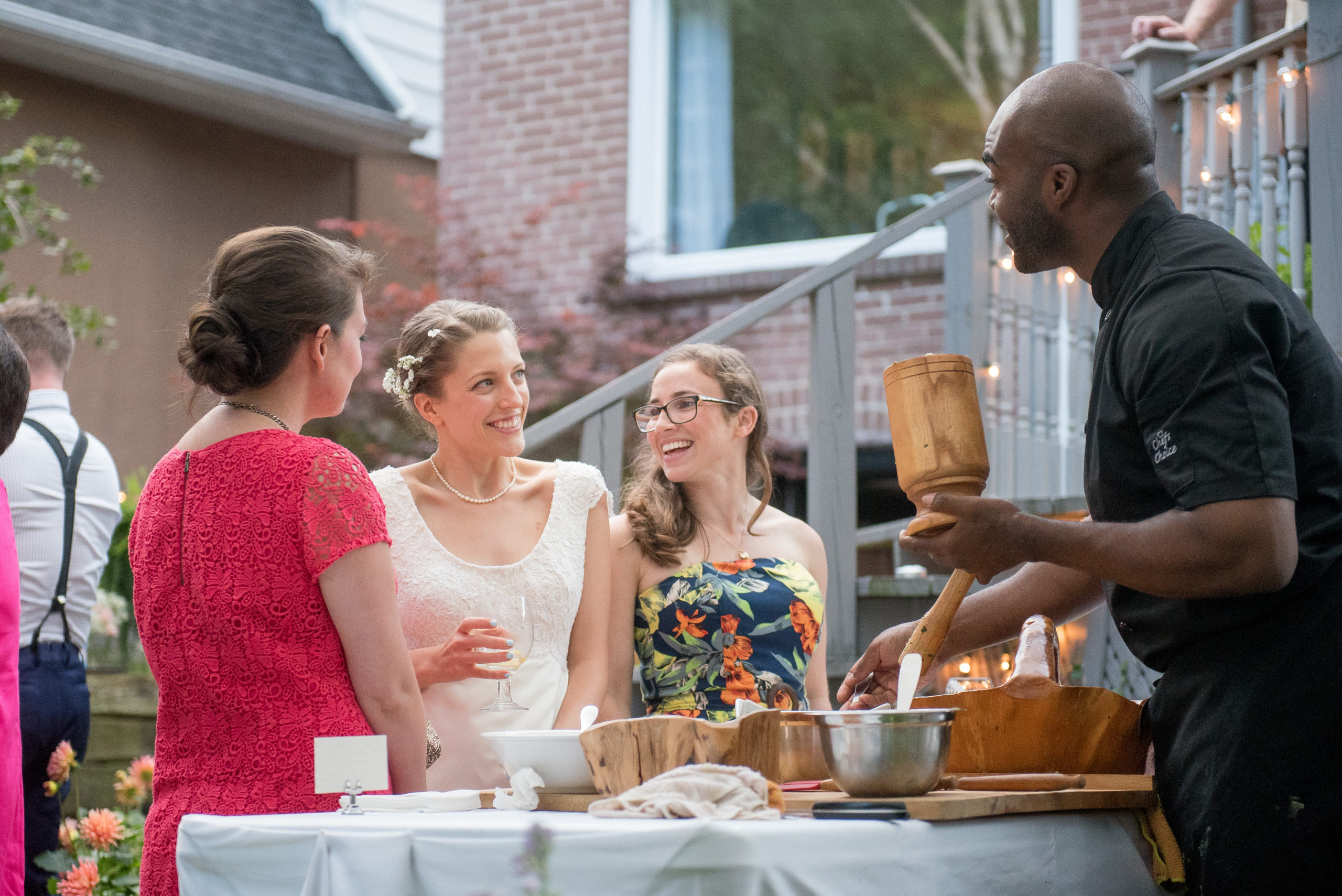 A chef serves food to three smiling women at an outdoor event. The women are dressed in colorful dresses and are gathered around a table, engaged in cheerful conversation. A brick house and greenery are in the background.