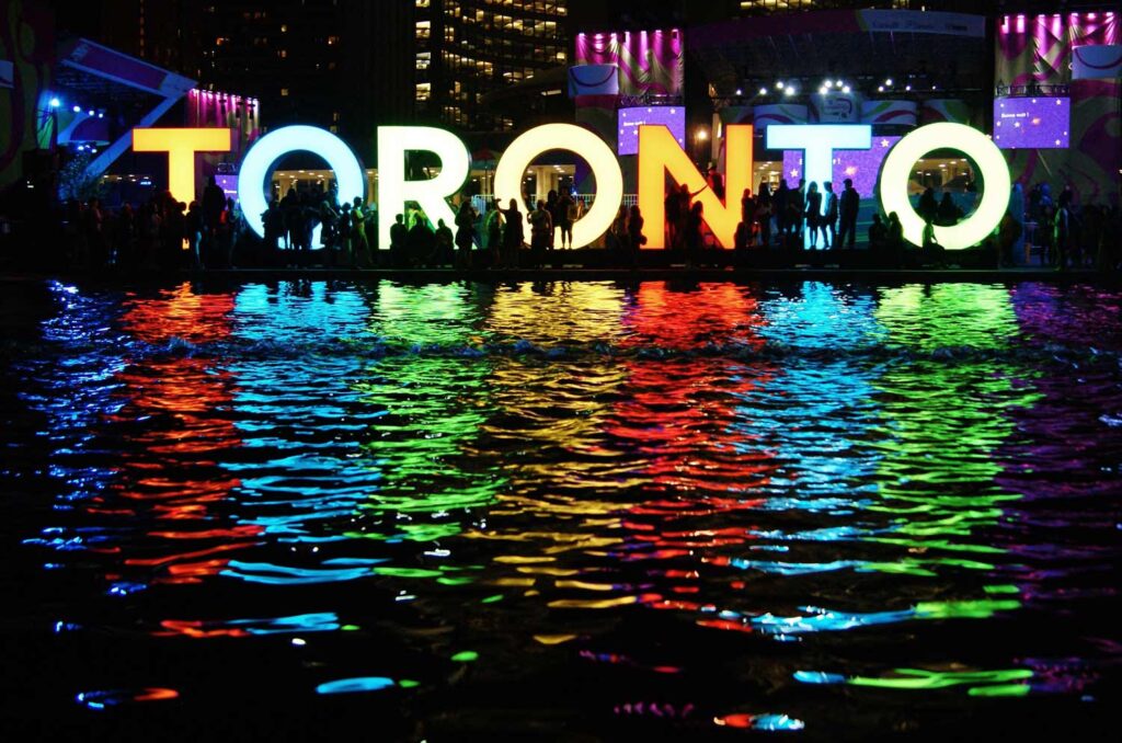 Large illuminated letters spelling "Toronto" reflected in colorful patterns on water at night, with silhouettes of people near Hall Crown and city lights glowing in the background.