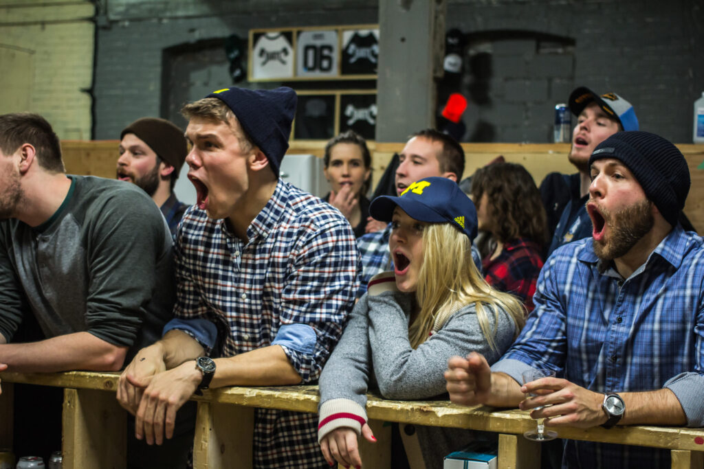 A group of enthusiastic people, some wearing plaid shirts and hats, cheer loudly behind a wooden barrier at an indoor BATL event, displaying excitement and surprise.
