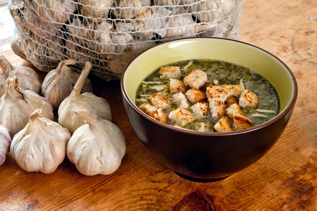 A bowl of green soup topped with croutons sits on a wooden table, accompanied by several garlic bulbs and a mesh bag brimming with more garlic.