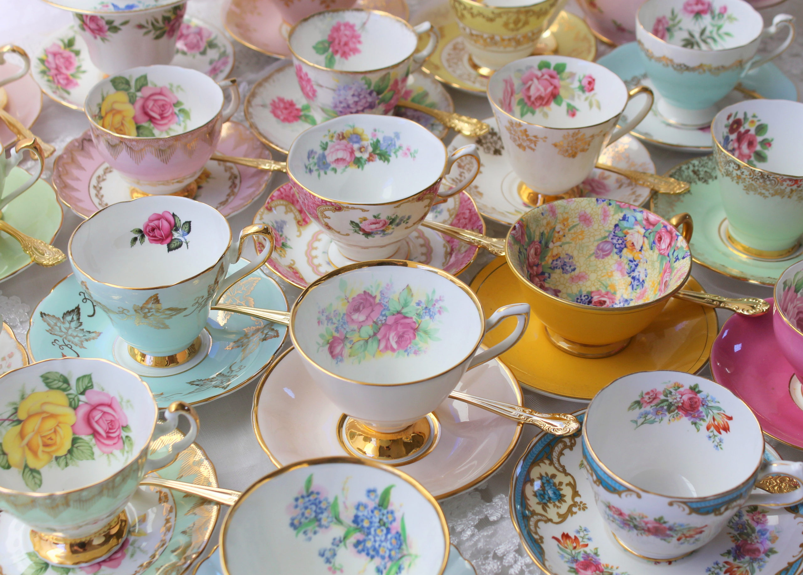 A collection of ornate, vintage teacups and saucers, each decorated with colorful floral patterns and gold trim, arranged closely together on a light surface—perfect for a charming afternoon tea shower.