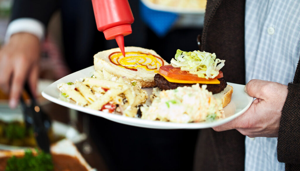 A person squeezes mustard onto a hamburger bun on a plate filled with a burger topped with cheese and veggies, crinkle-cut fries with ketchup, and a side of coleslaw.