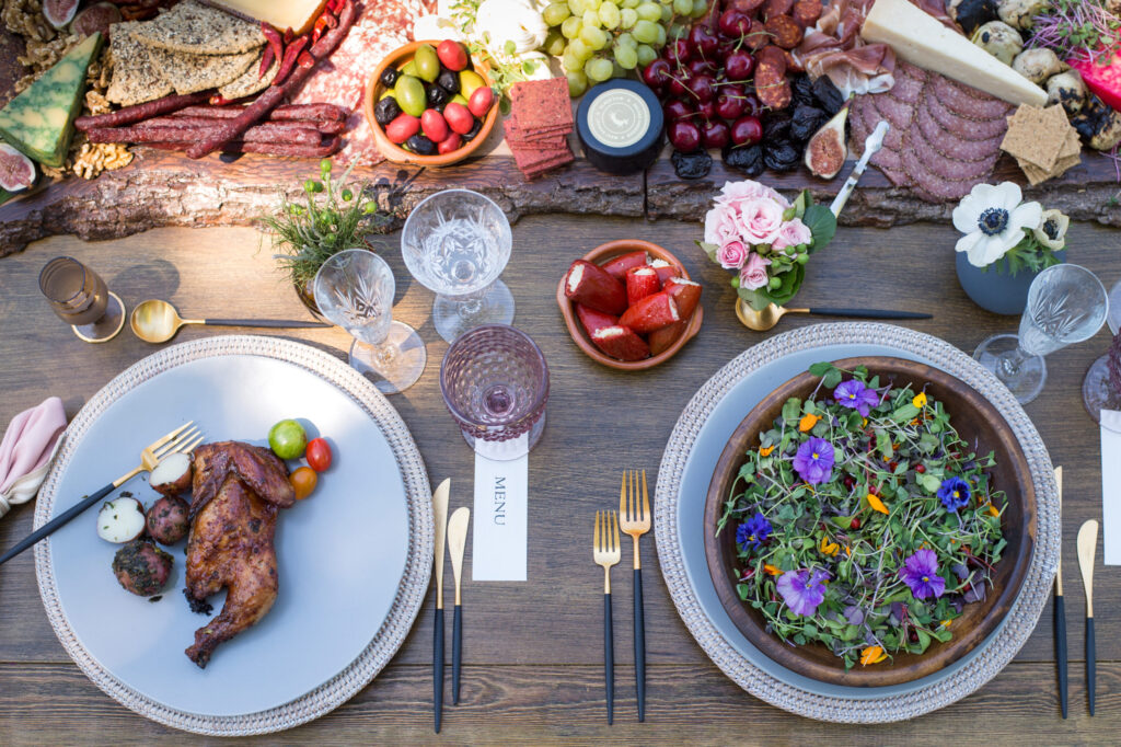 A beautifully set outdoor wedding table with roast chicken, colorful salad topped with edible flowers, fruit, charcuterie, cheeses, wine glasses, and floral decorations on a wooden surface.