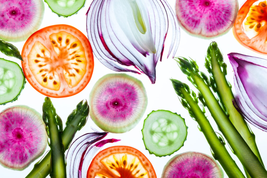 Sliced spring vegetables including asparagus, red onions, cucumbers, tomatoes, and watermelon radishes are arranged on a white background, showcasing their vibrant colors and fresh textures.