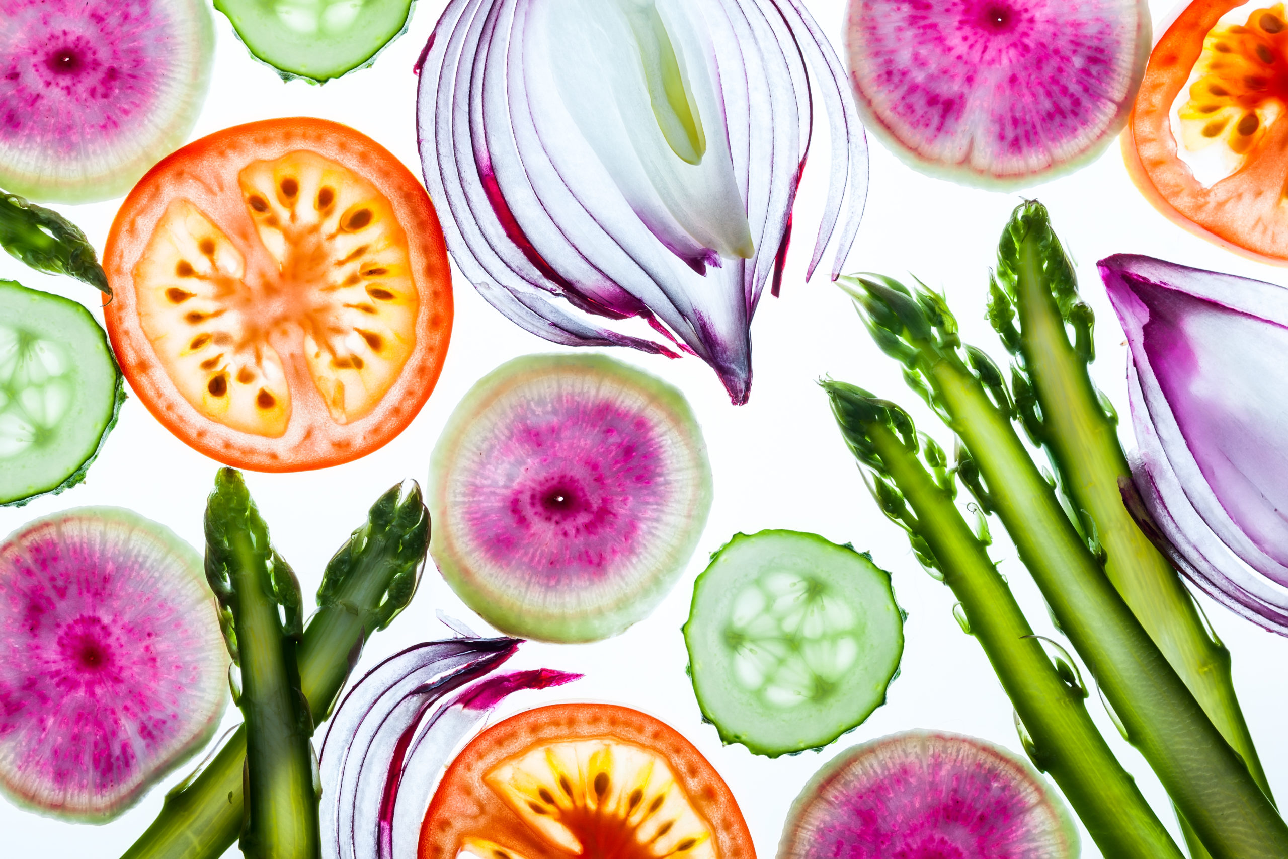 Sliced spring vegetables including asparagus, red onions, cucumbers, tomatoes, and watermelon radishes are arranged on a white background, showcasing their vibrant colors and fresh textures.