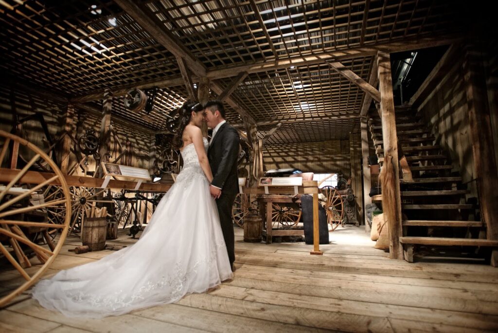 A bride and groom stand close together, facing each other and holding hands in a rustic wooden barn at Markham Museum, surrounded by old wagons and tools, with soft light filtering through the slatted ceiling.