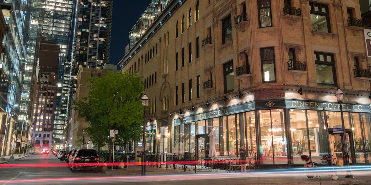 A lively city street at night with tall modern buildings, vibrant venues, a historic brick building housing Dineen Coffee Co., lit windows, parked cars, and light trails from passing vehicles.