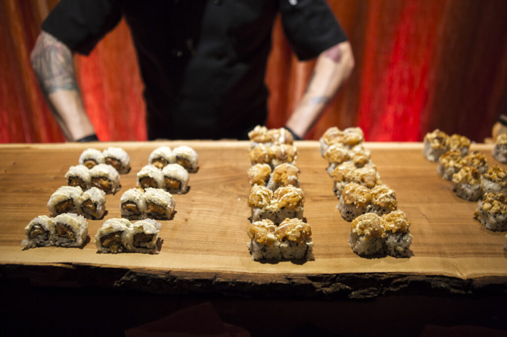 A chef stands behind a wooden board displaying three neat rows of sushi rolls with different toppings. The warmly lit, reddish background enhances the fresh sushi presentation.