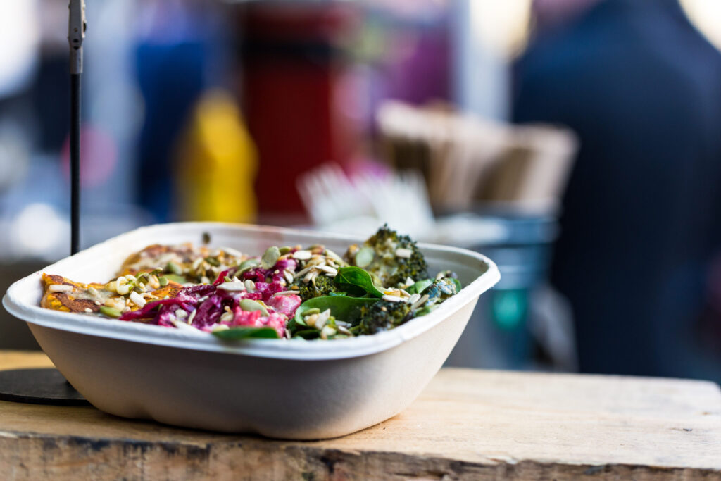 A compostable takeout container filled with a colorful salad, including greens, broccoli, and pickled onions, sits on a wooden surface with a blurred background of a food market.