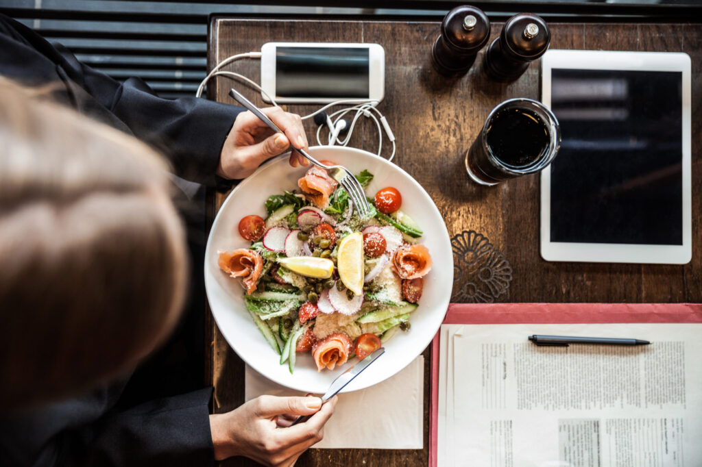 Person enjoying a salad with smoked salmon, radish, cherry tomatoes, and lemon at a wooden table set with corporate catering essentials—phone, earphones, tablet, drink, and documents. View is from above.