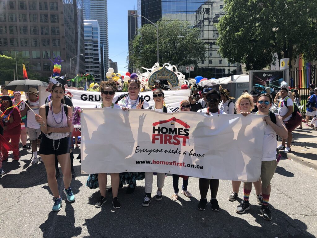 A diverse group of people stand outdoors, smiling and holding a large Homes First banner at a parade, surrounded by colorful balloons, festive outfits, and city buildings in the background.