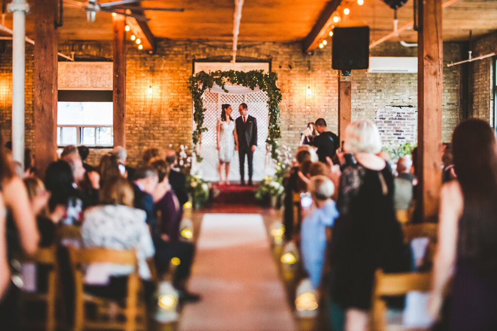 A bride and groom stand at the altar of the Jam Factory, a warmly lit indoor venue, surrounded by seated guests. The aisle is decorated with candles, and greenery adorns the arch behind the couple.