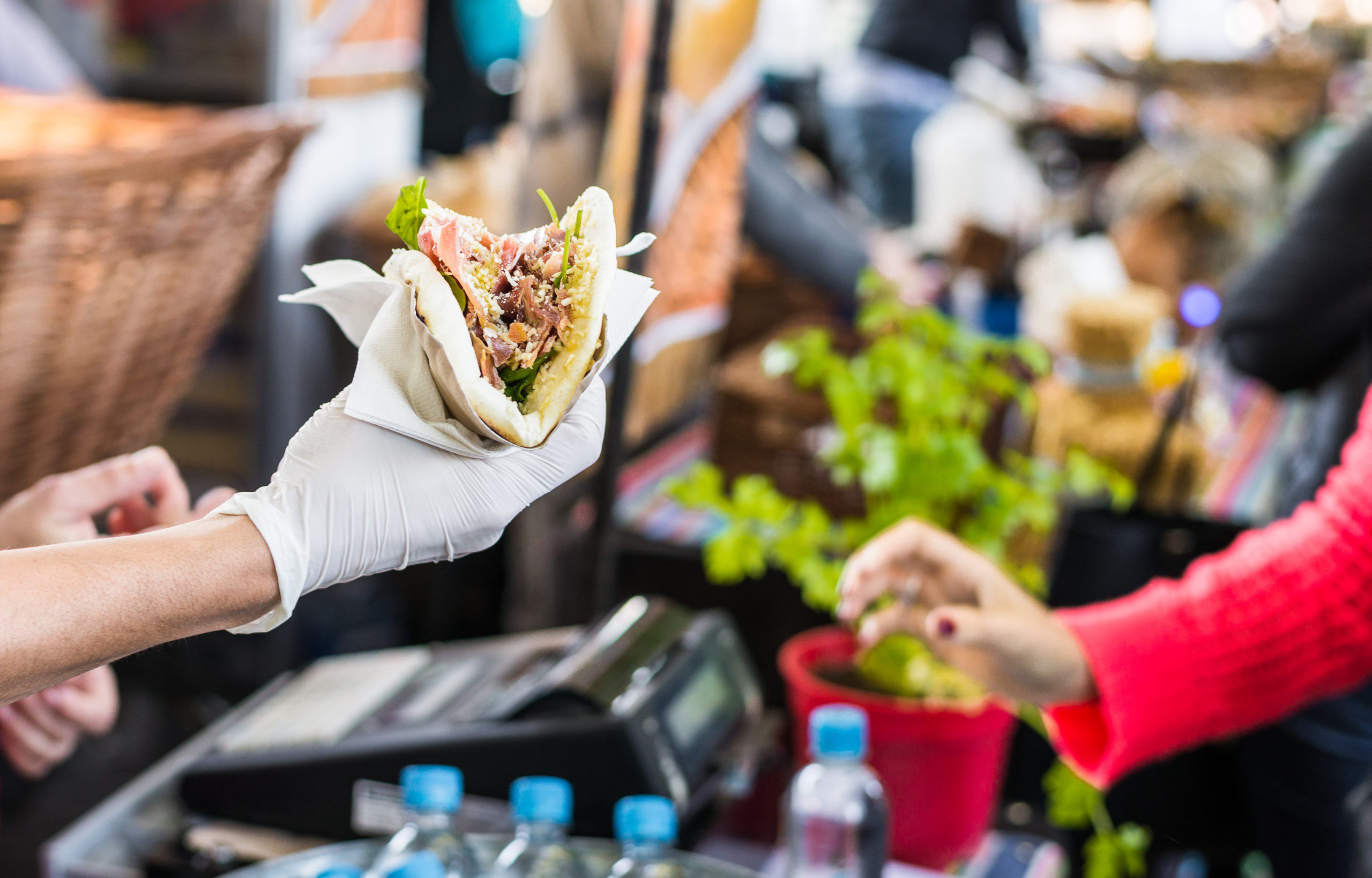 A gloved hand holds a filled wrap at an outdoor food market, perfect for corporate catering, while another person reaches out. Bottles of water, a potted herb, and baskets are visible in the background.
