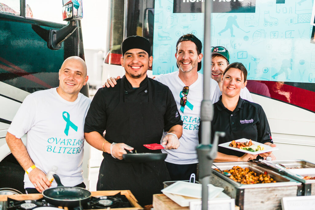Five people stand smiling at a food booth during Scotiabank Road Hockey to Conquer Cancer. Two wear "Ovarian Blitzer" shirts, one in a black chef uniform holds a spatula, and two others have food plates. Food trays are displayed in the foreground.