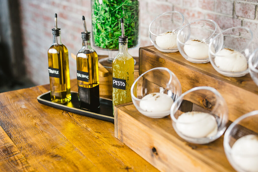 Bottles labeled truffle, sea spice, and pesto oil sit on a wooden table next to dishes of fresh mozzarella balls, with a jar of leafy greens and a rustic brick wall in the background.