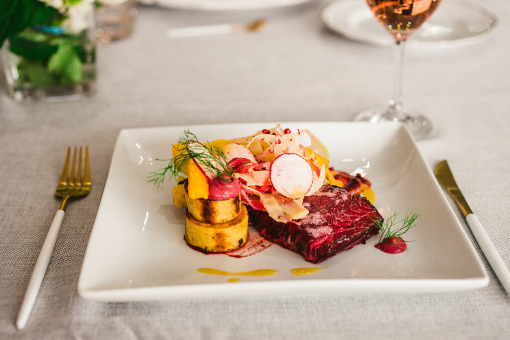 A gourmet dish is artfully plated with Valentine's Day Salmon, colorful vegetables, a rolled potato side, and herbs, served on a square white plate next to gold and white cutlery and a glass of rosé wine.
