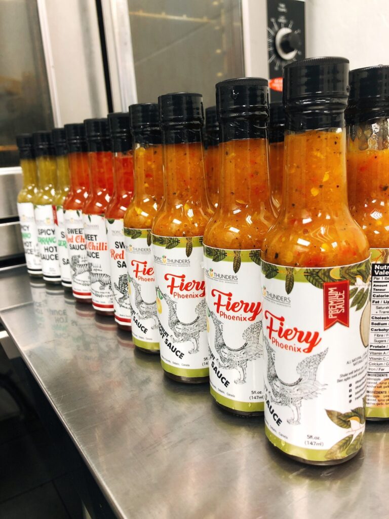 Several bottles of hot sauce, including Fiery Phoenix flavor, are lined up on a stainless steel countertop in a commercial kitchen at Pathways on Pleasure Valley. The sauces boast vibrant red, orange, and yellow hues.