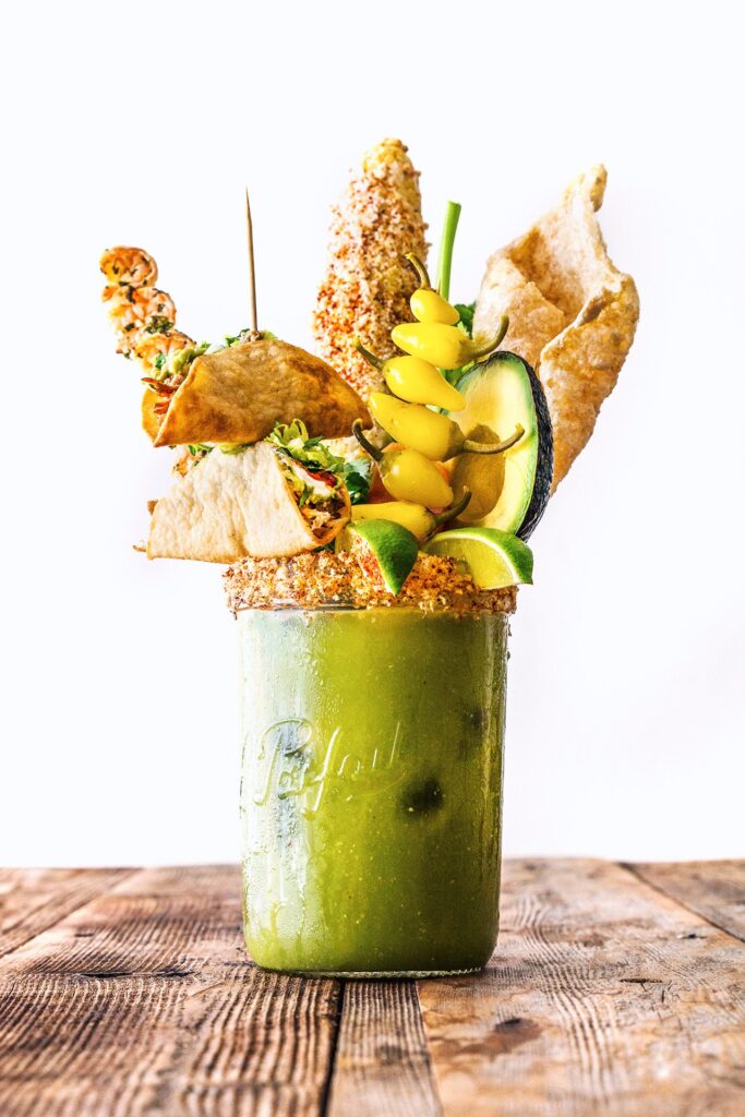 A green drink in a mason jar is topped with elaborate garnishes—taco shells, skewered yellow tomatoes, crispy chips, cucumber, and a lime wedge—perfect for festive Superbowl or Valentines Day Menus, set on a rustic wooden table with a white background.