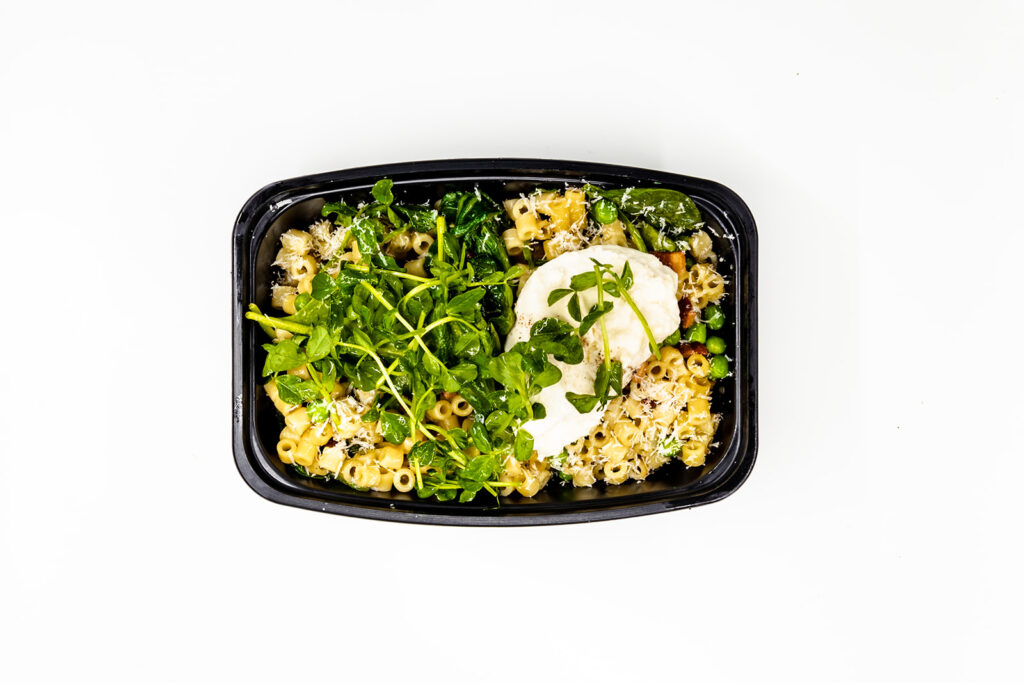 A black container filled with pasta, leafy greens, green peas, shredded cheese, and tender chicken breast slices, garnished with herbs—perfect for Superbowl and Valentines Day Menus, presented elegantly on a white background.