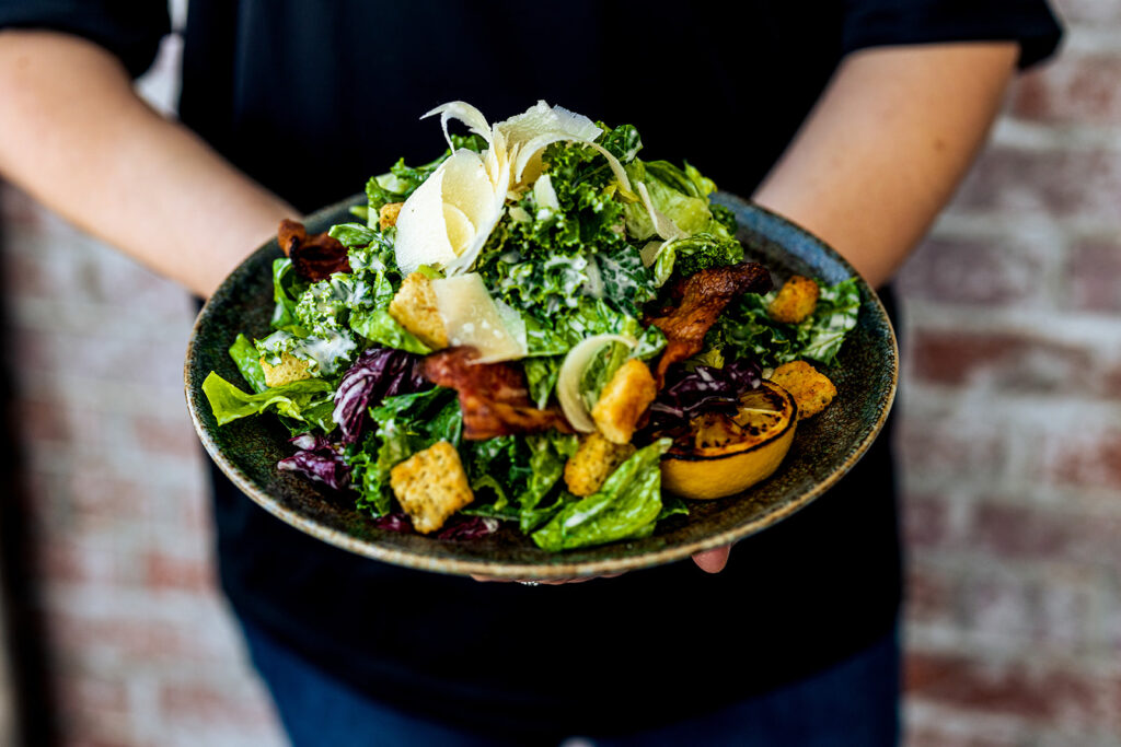 A person holds a large bowl of salad with leafy greens, croutons, crispy bacon, Parmesan cheese, and a grilled lemon half—perfect for Superbowl or Valentines Day menus—against a brick wall background.