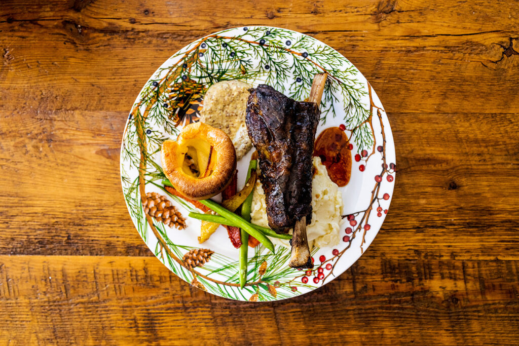 A decorated plate with braised meat on the bone, mashed potatoes, green beans, roasted carrots, Yorkshire pudding, and gravy—perfect for special occasions like Superbowl and Valentines Day menus—set on a rustic wooden table.