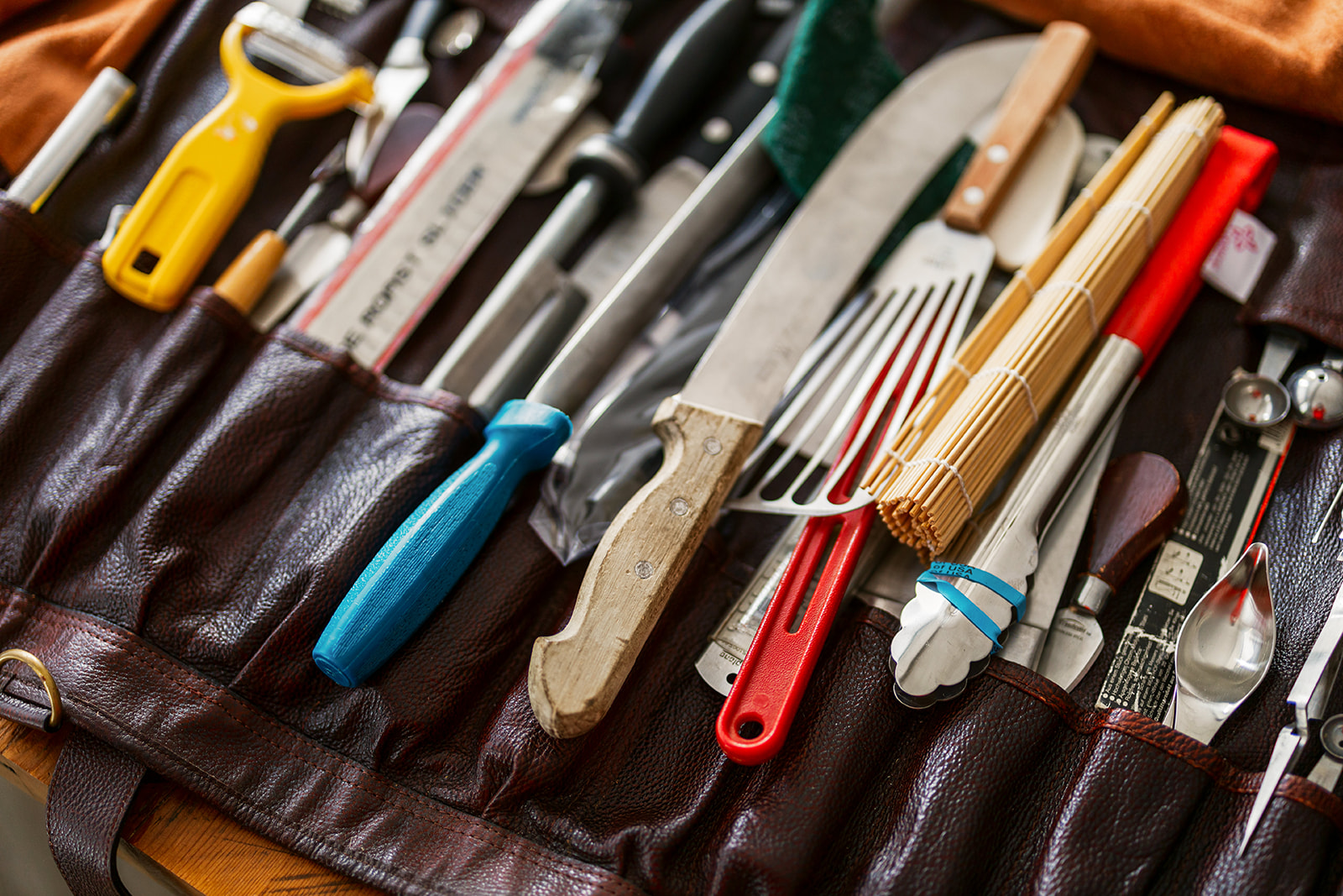 A close-up of a leather roll-up case filled with various kitchen tools, just like those used by an Executive Chef at Kiss the Cook Catering—featuring knives, a peeler, whisk, pastry brush, measuring spoons, bamboo sushi mat, and more.