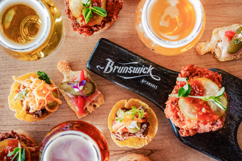 A wooden table features craft beers in glasses and assorted small appetizers on bread and crisps, served on a paddle with the Brunswick Bierworks logo—all arranged for an inviting Brunswick Bierworks tasting experience.