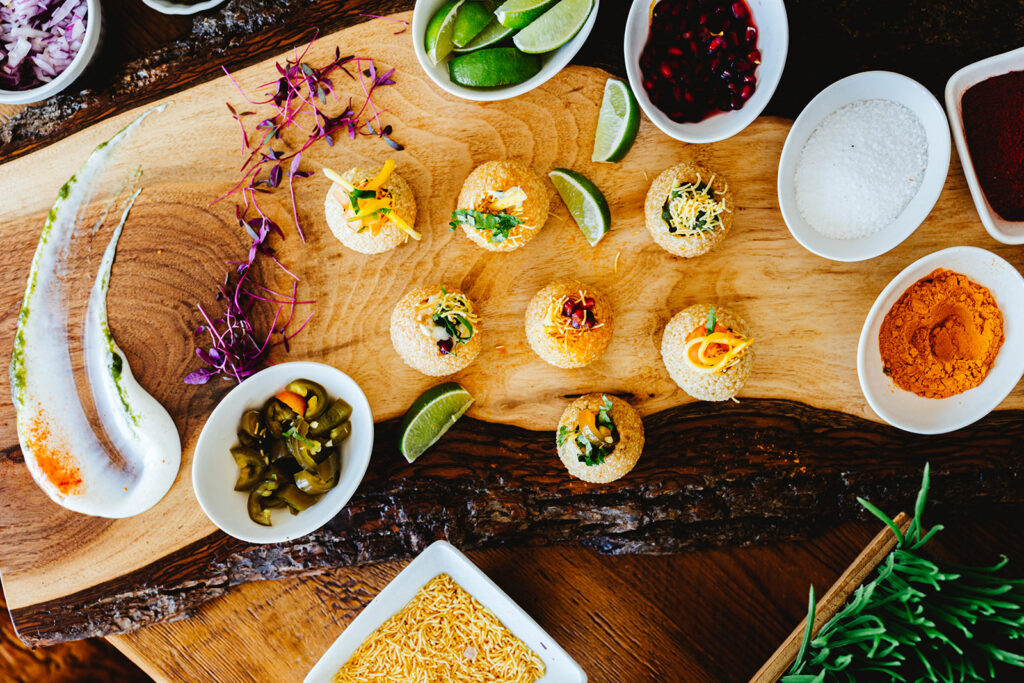 A wooden board from the hottest new food station, topped with assorted Indian snacks (pani puri), lime wedges, and colorful garnishes, surrounded by bowls of spices, chutneys, chopped onions, sev, and jalapeños.