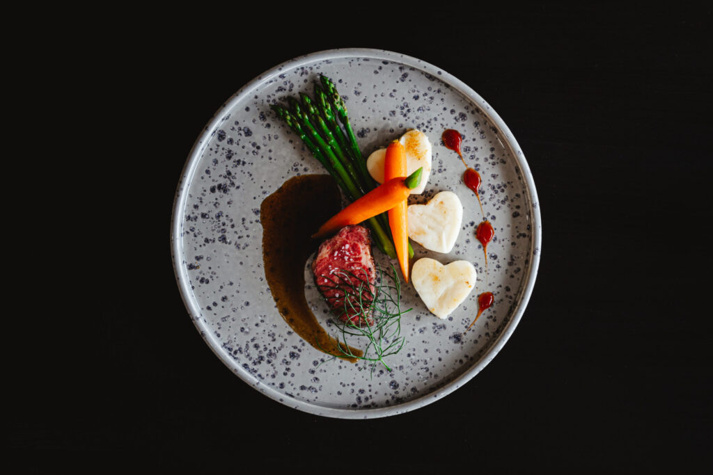 A gourmet dish on a speckled plate with asparagus, steak topped with herbs, carrots, and two heart-shaped mashed potatoes—one of the Top 5 Best Valentine's Day Dinner Ideas—arranged artistically against a black background.