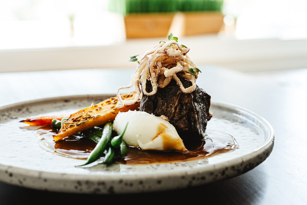 A gourmet dish featuring a piece of braised meat topped with crispy fried onions, served with mashed potatoes, green beans, and a golden slice of vegetable, all drizzled with brown sauce on a speckled plate.
