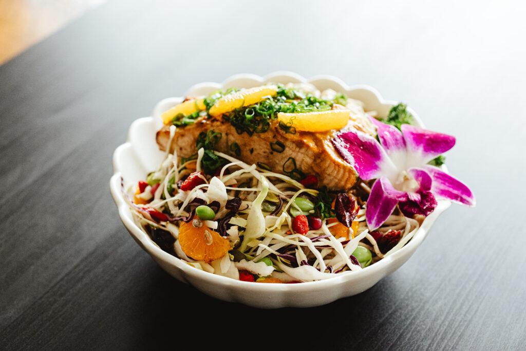 A white bowl filled with grilled salmon on top of a colorful salad with shredded vegetables, orange slices, edamame, seeds, and garnished with a purple orchid flower.