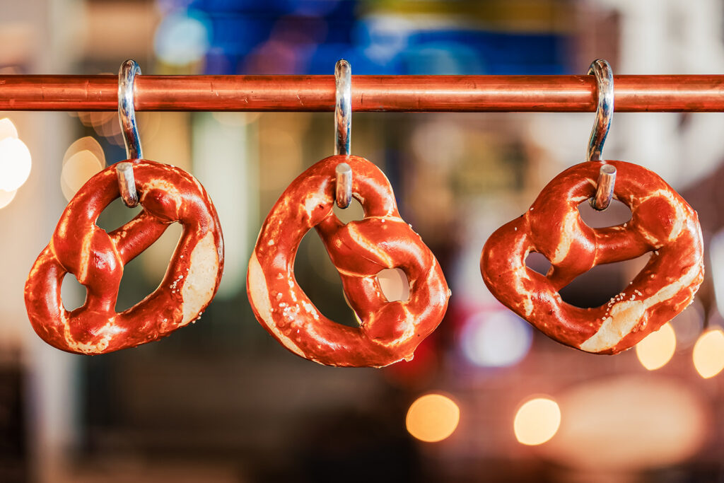 Three salted pretzels hang on metal hooks from a horizontal rod, with a blurred background of warm lights and colors, creating a festive and inviting atmosphere.