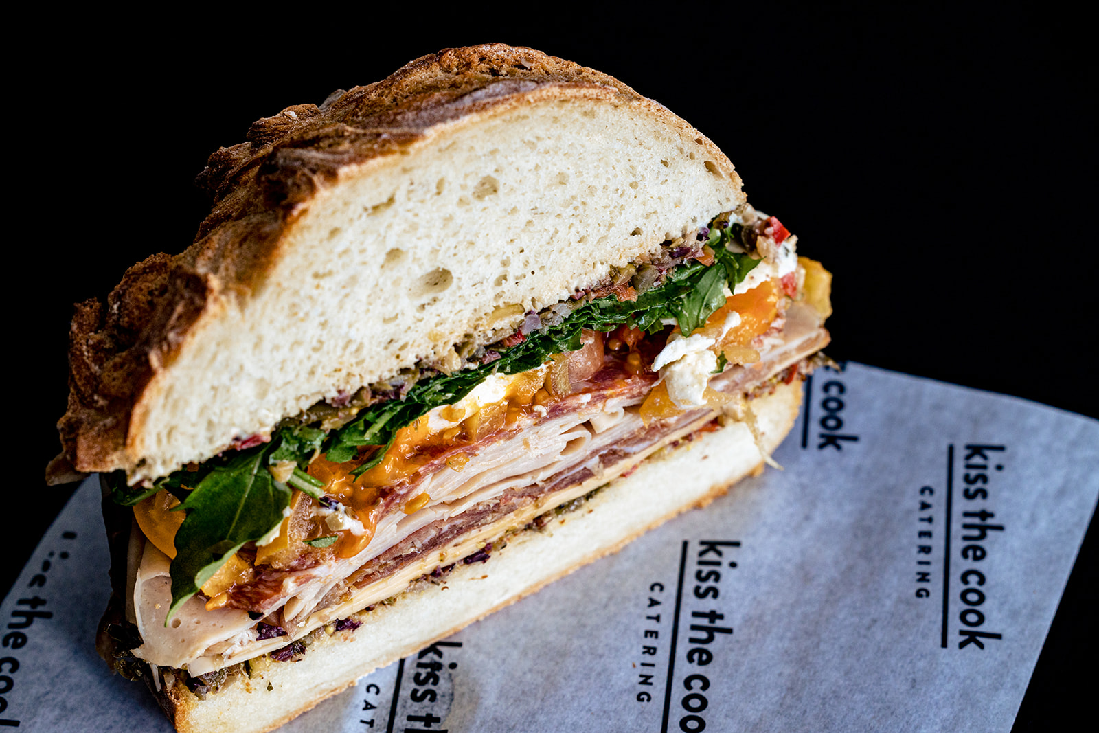 A thick sandwich on rustic bread, filled with turkey, cheese, bacon, and greens, sits on a sheet of "kiss the cook catering" paper—Corporate Picnic Perfection against a sleek black background.