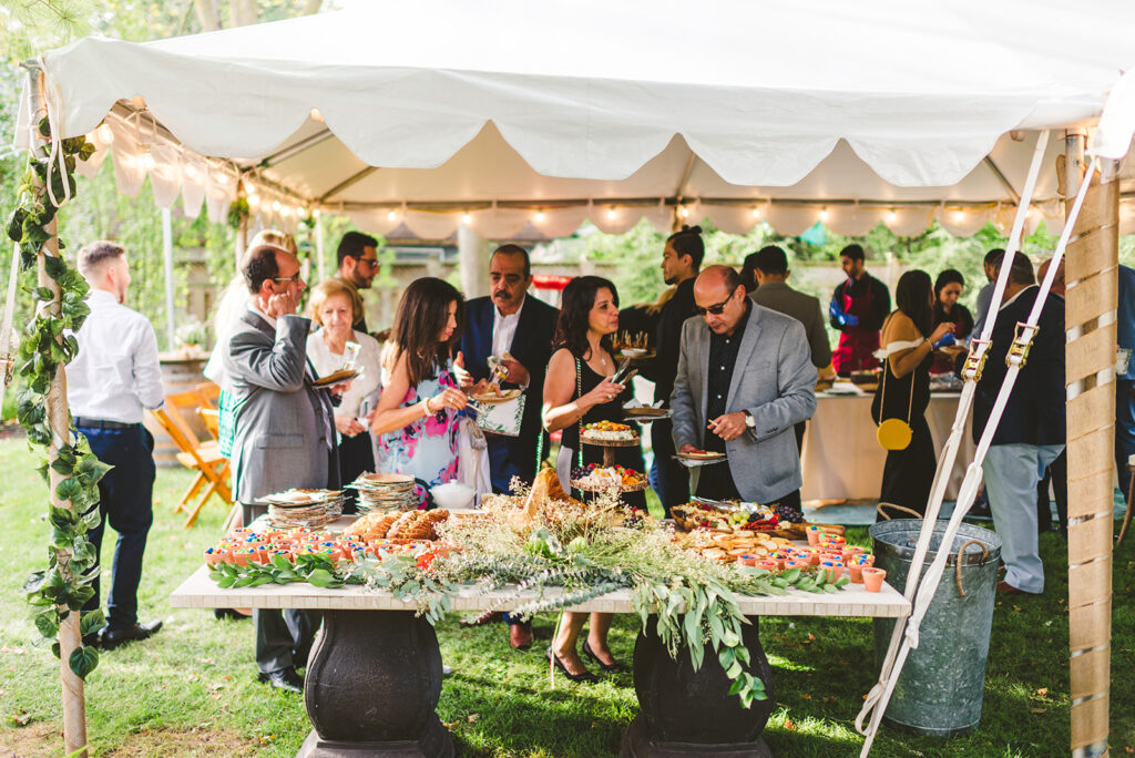 A group of people gather under a white tent outdoors, serving themselves food from a decorated buffet table filled with assorted dishes, greenery, and flowers at a daytime event.