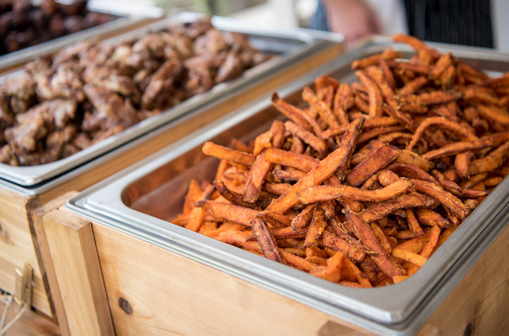 Two metal trays filled with food sit at a wooden buffet station, perfect for corporate catering. The front tray offers seasoned sweet potato fries, while the tray behind features flavorful cooked chicken wings.