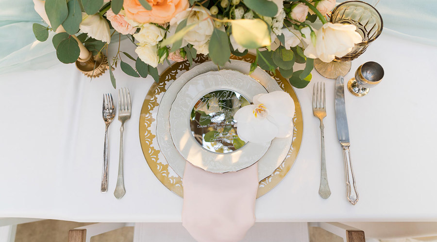 Elegant table setting with layered white and gold plates, a pink napkin, silver cutlery, a clear menu, a gold-rimmed glass, and a centerpiece of peach and white flowers. Perfect for any Toronto catering event. An orchid rests on the plate.