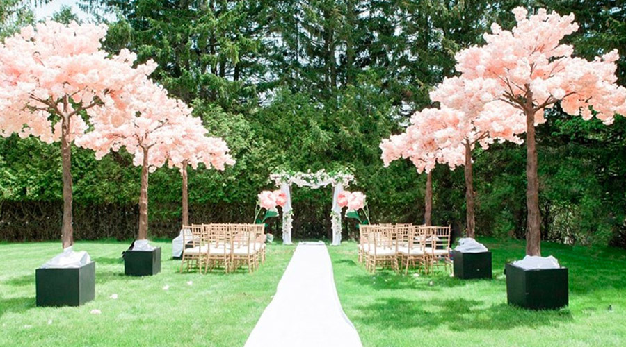 An outdoor wedding setup features a white aisle runner, rows of gold chairs, and large artificial cherry blossom trees with pink flowers, set against a backdrop of lush green trees—perfect for Toronto catering events.