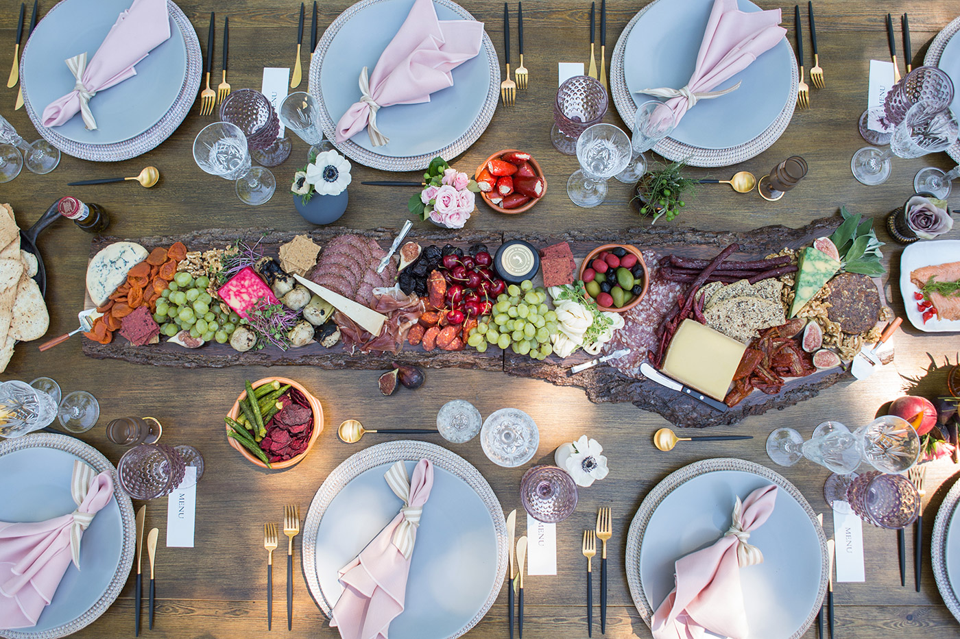 A wooden table set for a meal with pale blue plates, pink napkins, gold cutlery, and crystal glasses. Perfect for corporate catering, a colorful charcuterie board with fruits, cheese, crackers, and flowers runs down the center.
