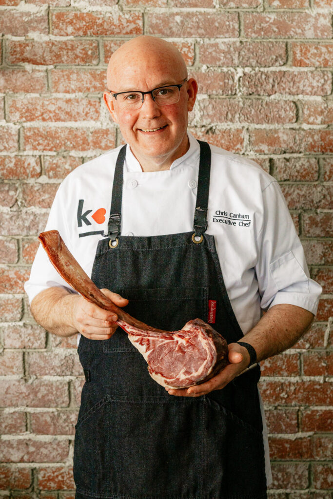 A smiling chef from a Toronto catering company in a white coat and black apron stands against a brick wall, proudly holding a large raw tomahawk steak.