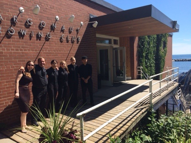 Six people dressed in black stand and smile outside the Argonaut Rowing Club building, which has a brick wall, ramp, and a view of blue sky and water in the background.