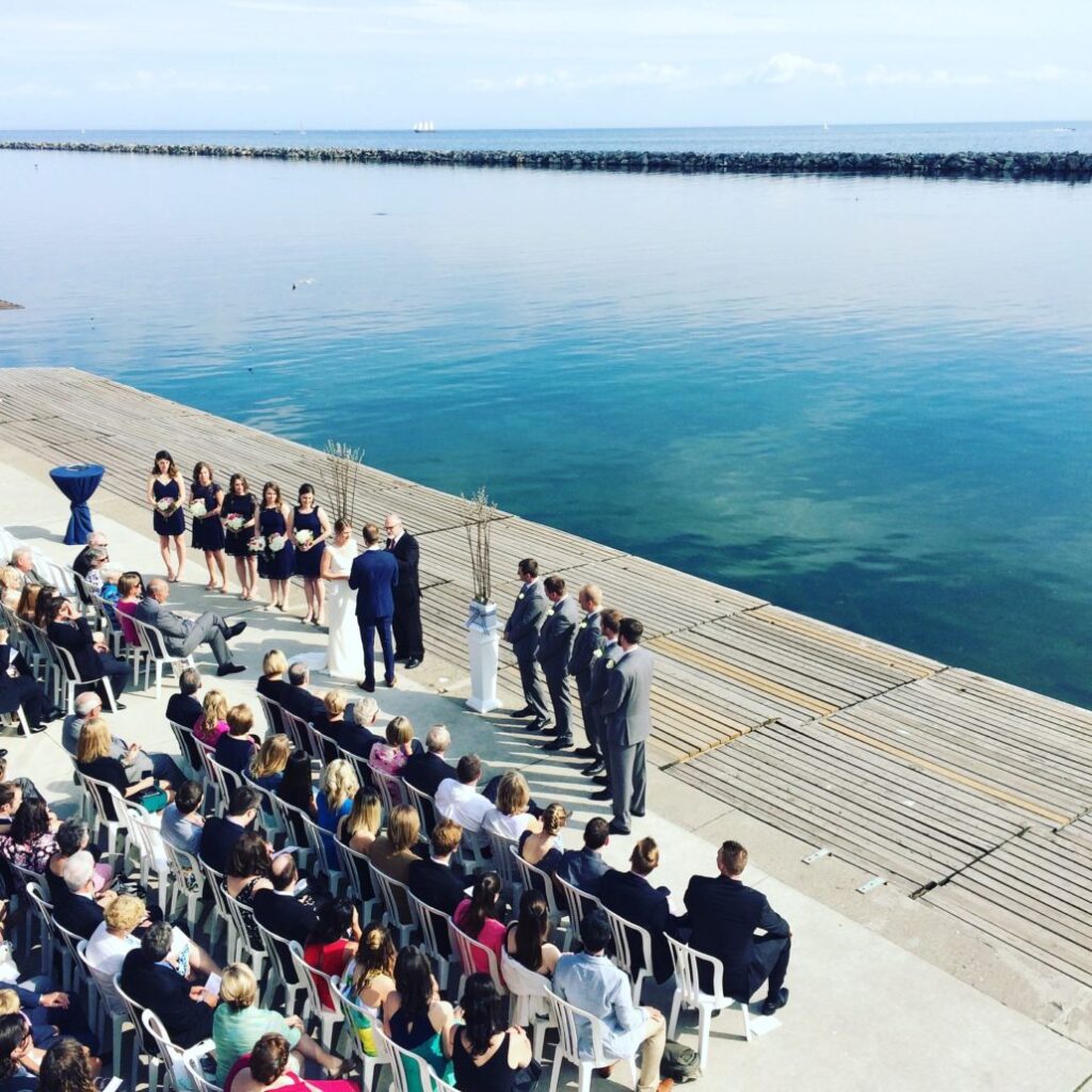 A wedding ceremony takes place outdoors beside a calm body of water, with guests seated on white chairs, the wedding party standing at the front, and blue sky reflected in the water.