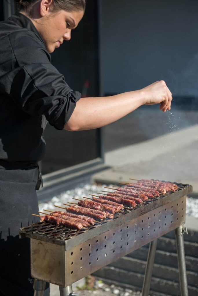 A person wearing a black outfit sprinkles seasoning onto skewers of meat grilling on an outdoor barbecue, with sunlight casting shadows on the scene.