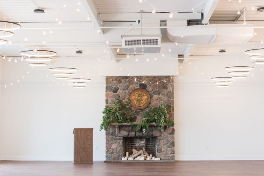 A stone fireplace decorated with green plants and candles is centered against a white wall in a bright room. A wooden podium stands to the left, and string lights are hung across the ceiling.