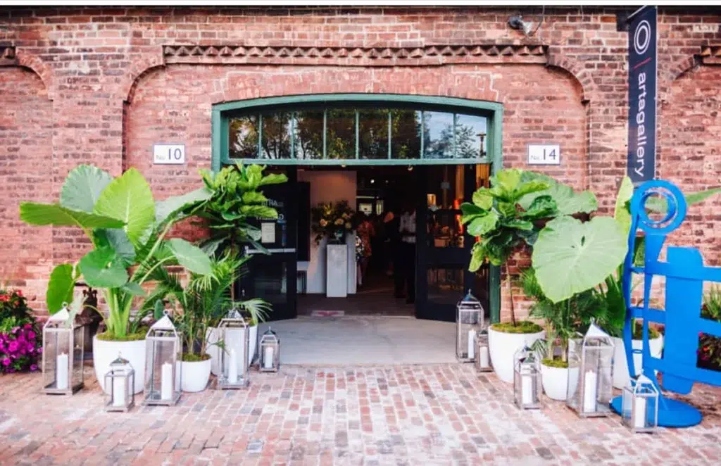 The entrance of a brick building with large glass doors, flanked by vibrant green potted plants and silver lanterns, leading into an art gallery. A banner with “gallery” is visible on the right.