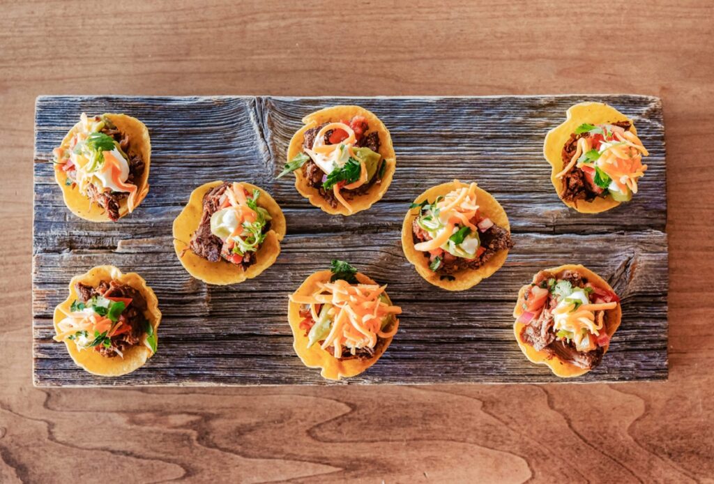Eight small tacos topped with shredded cheese, meat, vegetables, and sauce are arranged on a rustic wooden serving board—perfect for your next Corporate BBQ—set on a brown wooden table.