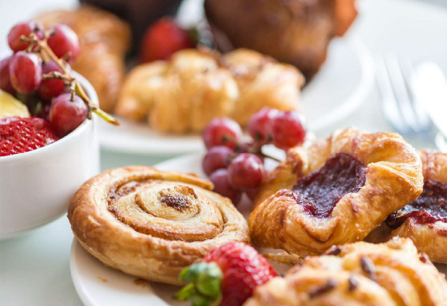 A close-up of assorted pastries, including a cinnamon roll and a jam-filled Danish, on a plate with fresh strawberries and grapes—perfect for event catering. A bowl of fruit and more pastries are blurred in the background.