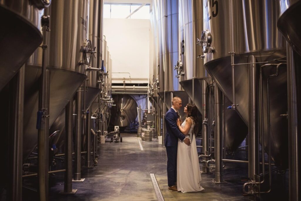 A couple in wedding attire stands closely together in the center aisle of a brewery, surrounded by large stainless steel brewing tanks. Natural light shines from windows above.