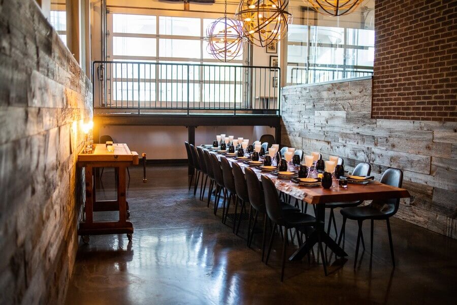A long dining table with black chairs is set for a meal in a modern, rustic room with wooden walls, large windows, and warm lighting from globe chandeliers.