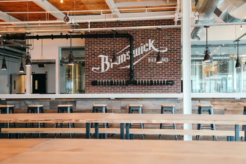Spacious brewery taproom with wooden tables, industrial lighting, and a brick wall featuring the "Brunswick Bierworks" logo. High stools line the bar, and large windows reveal brewing equipment in the background.